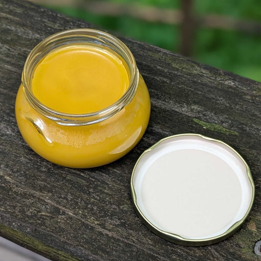 A small clear glass jar filled with bright yellow ghee or butter, placed on a weathered wooden surface with its white lid set beside it.