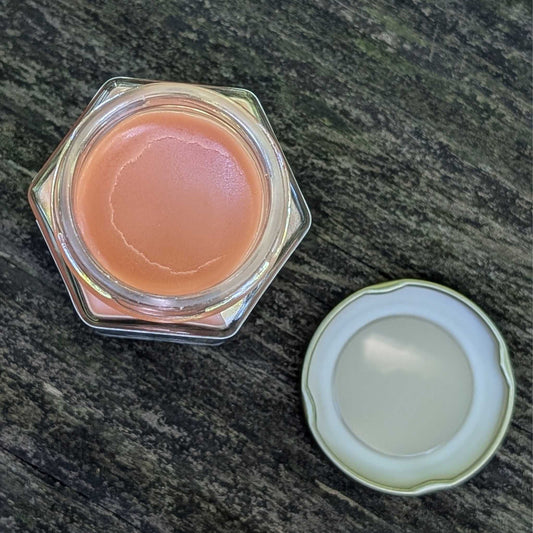 A hexagonal clear cosmetic jar filled with peachy pink salve shown open next to its white scalloped lid on a dark textured surface.