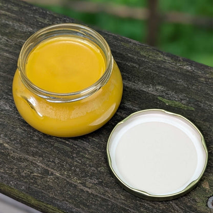 A small clear glass jar filled with bright yellow ghee or butter, placed on a weathered wooden surface with its white lid set beside it.
