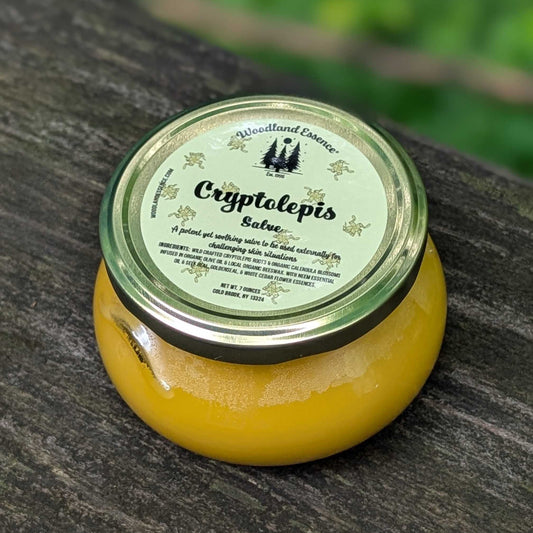 A glass jar of Cryptolepis Salve by Woodland Essence, filled with a yellow balm and sealed with a green glass lid and a white label, resting on a dark weathered wooden surface.