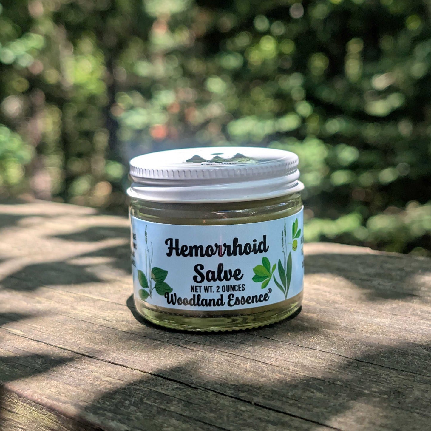 A small glass jar of Woodland Essence Hemorrhoid Salve with a white lid and white label, sitting on a wooden surface with a blurred green outdoor background.