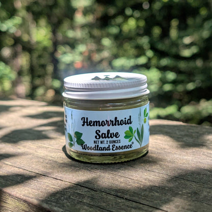 A small glass jar of Woodland Essence Hemorrhoid Salve with a white lid and white label, sitting on a wooden surface with a blurred green outdoor background.